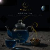 Chaleira e xícara transparentes contendo chá azul da marca Mystea sobre superfície que lembra o solo lunar, com um pequeno recipiente de ervas ao lado e cenário espacial ao fundo com a Terra visível.