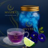 Jarro de chá azul gelado da Mystea com brilhos mágicos, ao lado de uma xícara de chá roxo, flores de Clitoria ternatea e uma fatia de limão sobre fundo azul escuro.
