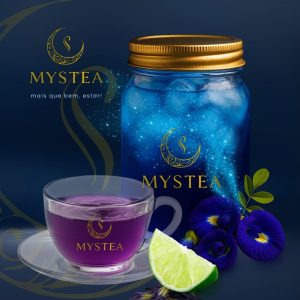 Jarro de chá azul gelado da Mystea com brilhos mágicos, ao lado de uma xícara de chá roxo, flores de Clitoria ternatea e uma fatia de limão sobre fundo azul escuro.