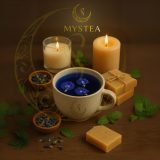 Xícara clara com chá azul decorado com flores de Clitoria ternatea, rodeada por velas acesas, folhas verdes, pequenas tigelas com pétalas secas e barras de sabonete artesanal, sobre fundo marrom com o logotipo Mystea ao fundo.