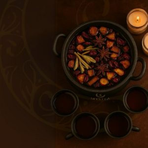 Vista superior de um caldeirão escuro contendo infusão vermelha com frutas, anis-estrelado e especiarias, acompanhado por três xícaras servidas e duas velas acesas sobre fundo marrom decorado.